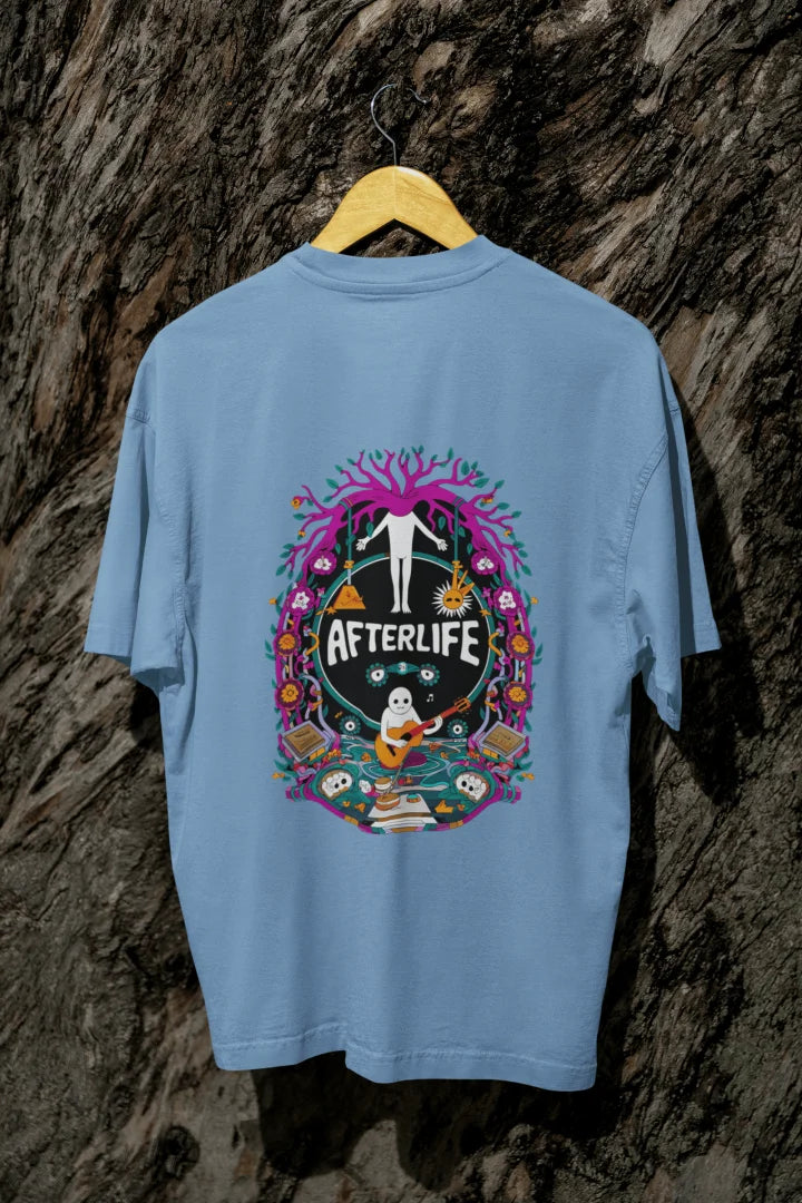 Blue oversized tee shirt hanging on a wooden hanger against a textured background, featuring a colorful psychedelic circular design with the word 'Afterlife' at the top. The design includes a tree with pink and purple leaves, and a skull and crossbones, surrounded by vibrant, intricate details.