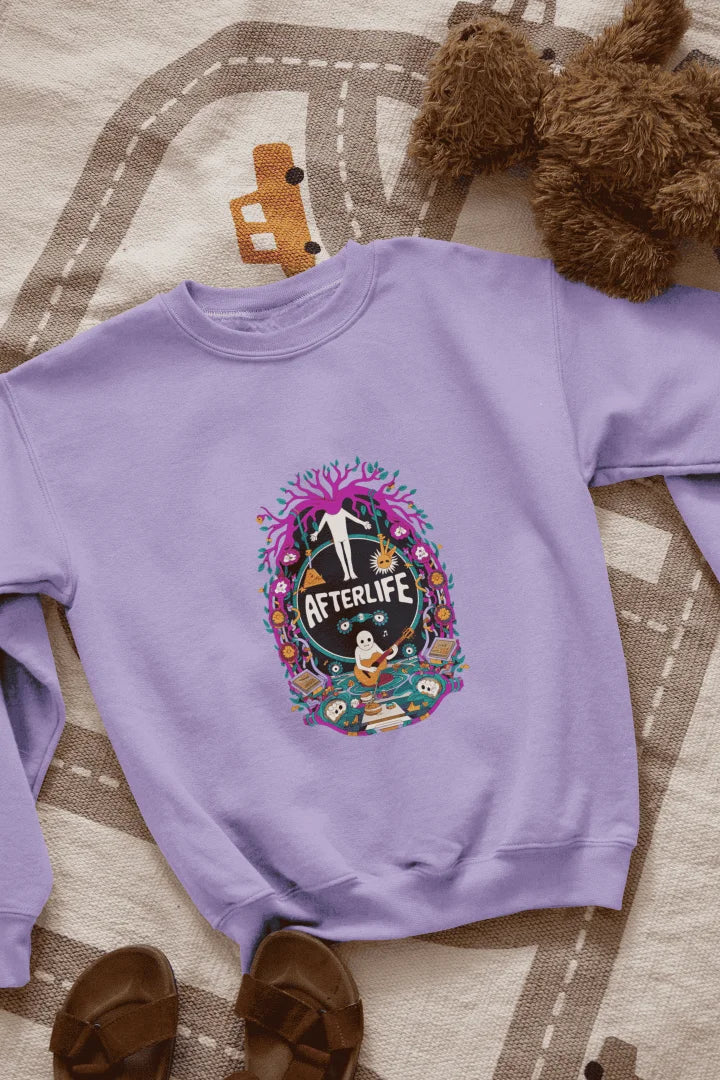 Lavender sweatshirt featuring a colorful graphic design with a circular Afterlife text, showing a person with outstretched arms standing in front of a tree with pink and purple leaves. The design includes a light blue sky with white clouds, a person in a blue shirt smiling, and brown sandals and a teddy bear on a beige and white patterned blanket.