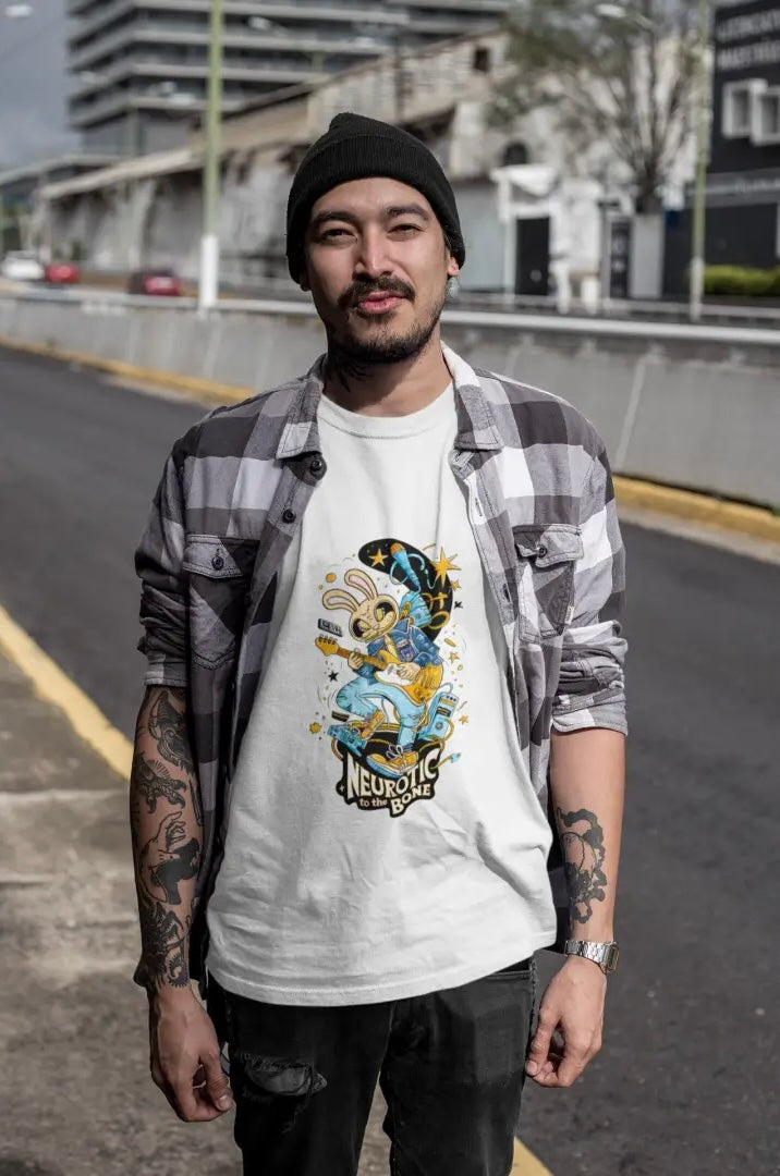 Young man wearing a white band T-shirt with a character playing guitar, black jeans, and a black beanie, standing on an empty street with a building and a tree in the background.