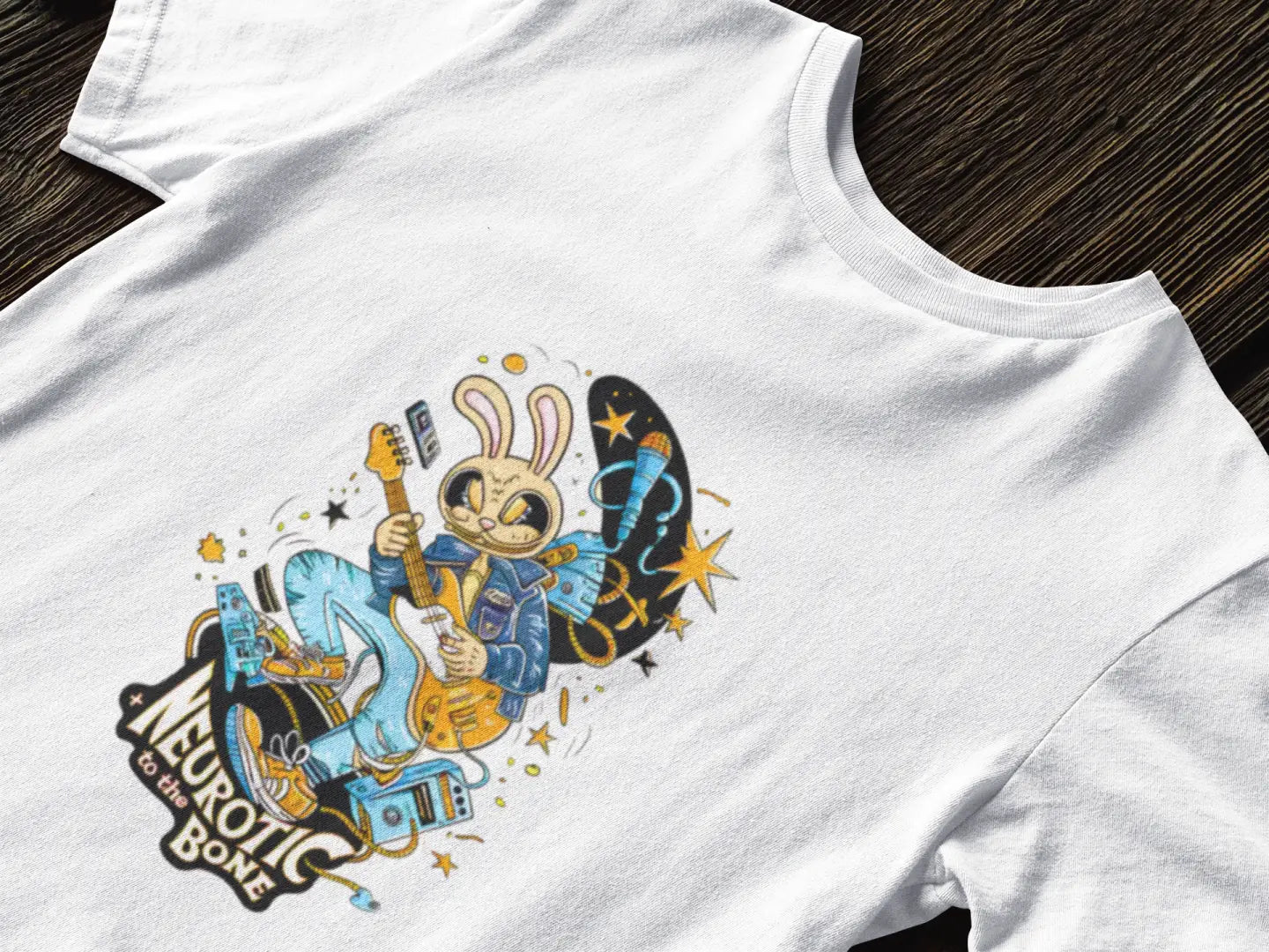 White band T-shirt featuring a playful illustration of a cartoon bunny in a blue suit, smiling and playing a guitar with both hands. The design includes colorful stars and musical notes in the background, with a banner above the bunny reading Neurotic To The Bone in black letters.