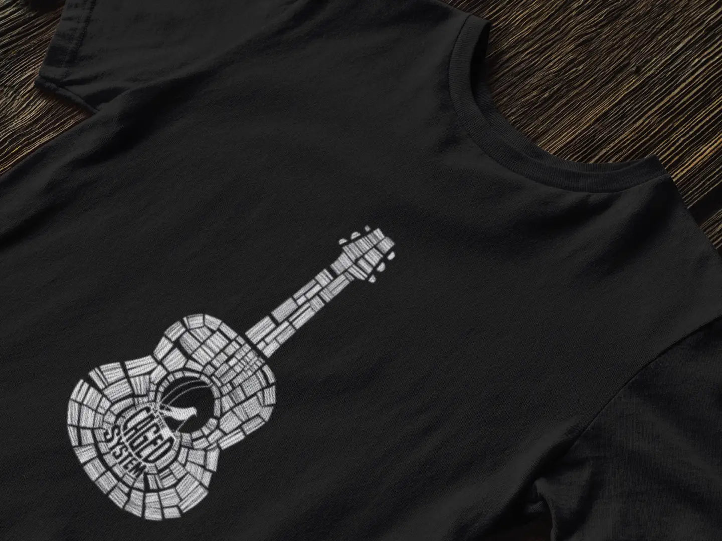 Black T-shirt with a graphic design of an acoustic guitar made up of small squares in a mosaic pattern, black neck, and visible strings. The design is set against a black background, laid flat on a wooden surface.