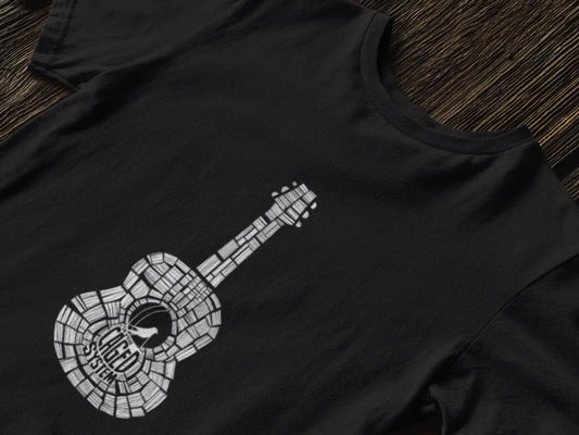 Black T-shirt with a graphic design of an acoustic guitar made up of small squares in a mosaic pattern, black neck, and visible strings. The design is set against a black background, laid flat on a wooden surface.
