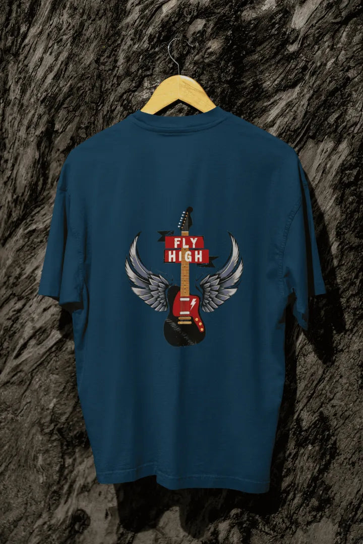 A navy blue oversized t-shirt with a design printed on the back hanging on a tree trunk. The design contains a guitar with wings. There's a text saying Fly High at the center top of the design.