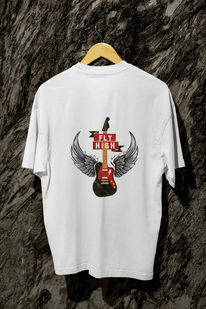 A white oversized t-shirt with a design printed on the back hanging on a tree trunk. The design contains a guitar with wings. There's a text saying Fly High at the center top of the design.