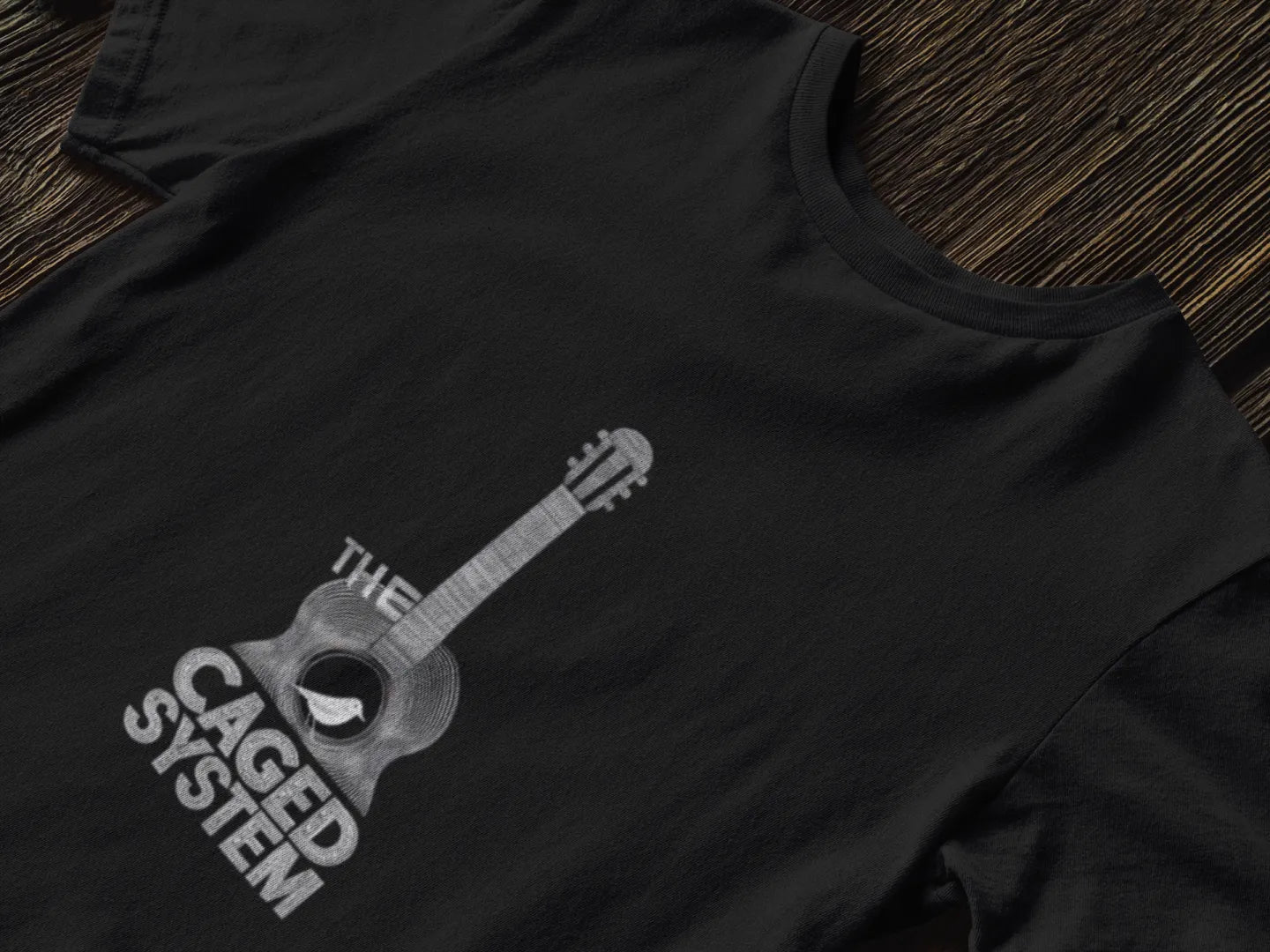A black T-shirt with a minimal design lying down on a table. The design consists of an acoustic guitar. A small bird is sitting on the hole of the guitar. There’s a text saying The Caged System.