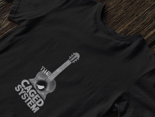 A black T-shirt with a minimal design lying down on a table. The design consists of an acoustic guitar. A small bird is sitting on the hole of the guitar. There’s a text saying The Caged System.