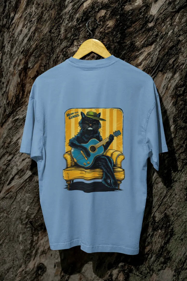A baby blue back printed oversized t-shirt with a back print hanging on a tree trunk. The print design contains a cat wearing a hat and sitting on the sofa. There's a text saying Meowsical Genius on the top left of the design.