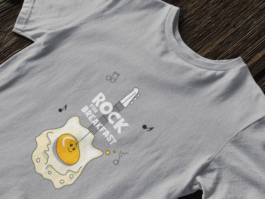 Close up of a grey t-shirt kept on a wooden floor with a design printed on the front. The design consists of a a guitar which appears to be like an egg. There are musical notations floating around the design. There's a text saying Rock For Breakfast at the center of the design.