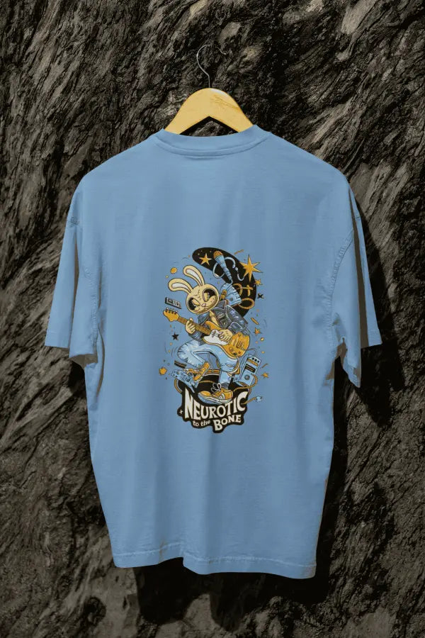 A baby blue oversized t-shirt with a design printed at the back hanging on a tree trunk. The design consists of a character playing a guitar. The overall design is funky. There's a text saying Neurotic to the Bone at the bottom of the design.