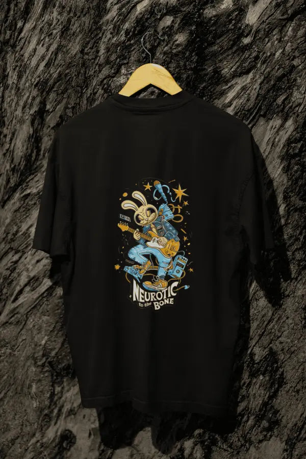A black oversized t-shirt with a design printed at the back hanging on a tree trunk. The design consists of a character playing a guitar. The overall design is funky. There's a text saying Neurotic to the Bone at the bottom of the design.
