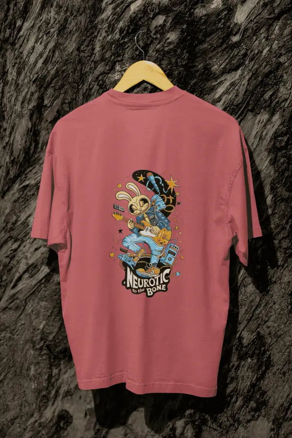 A dusty rose oversized t-shirt with a design printed at the back hanging on a tree trunk. The design consists of a character playing a guitar. The overall design is funky. There's a text saying Neurotic to the Bone at the bottom of the design.