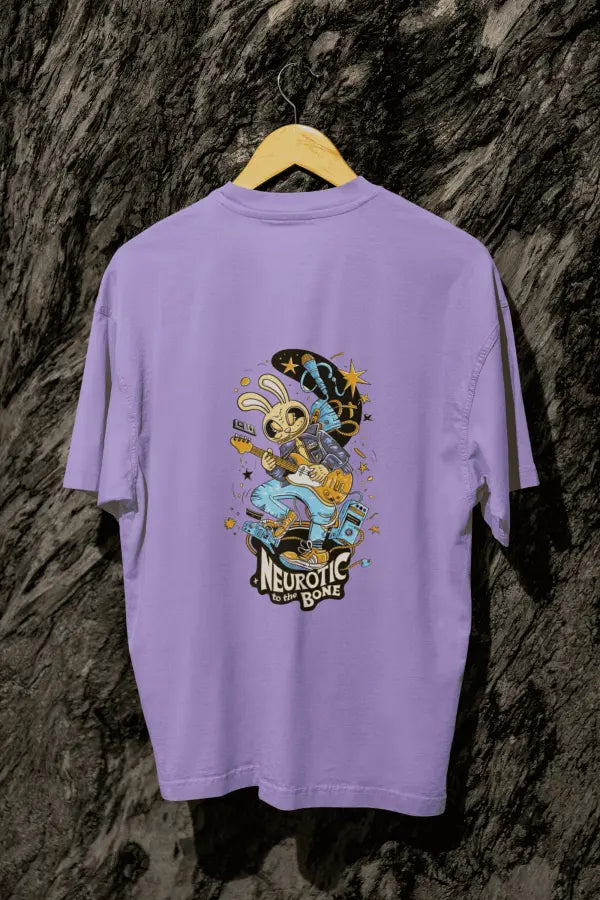 A lavender oversized t-shirt with a design printed at the back hanging on a tree trunk. The design consists of a character playing a guitar. The overall design is funky. There's a text saying Neurotic to the Bone at the bottom of the design.