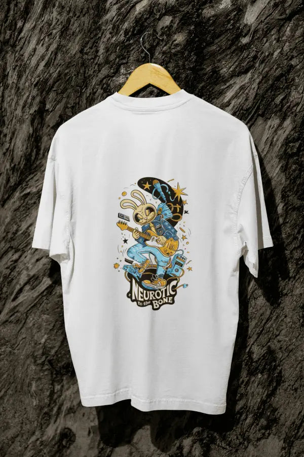 A white oversized t-shirt with a design printed at the back hanging on a tree trunk. The design consists of a character playing a guitar. The overall design is funky. There's a text saying Neurotic to the Bone at the bottom of the design.