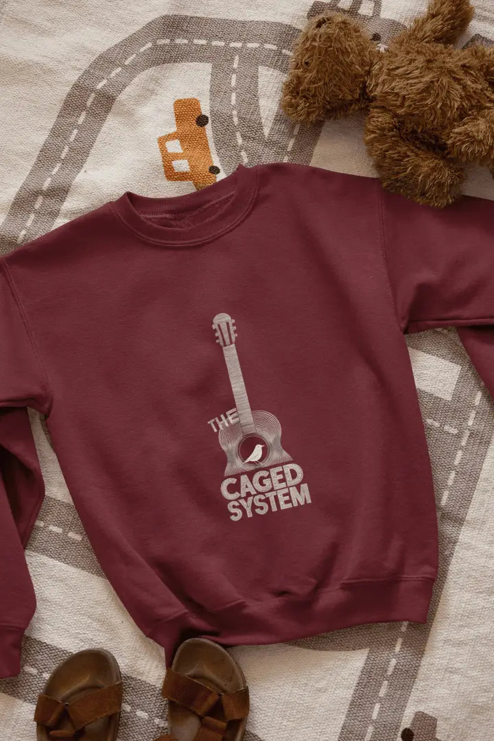 A maroon sweatshirt with a minimal design printed on the front lying on the floor. The design consists of an acoustic guitar inside which a small bird is sitting. There's a text saying The Caged System at the bottom of the design.