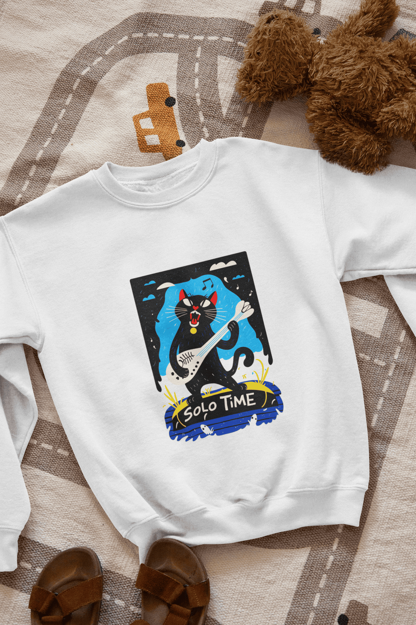A white sweatshirt with a design printed on the front lying down on the floor. The design contains a cat holding a fish bone like a guitar and appearing to play it aggressively. There's a text saying Solo Time at the bottom of the design.