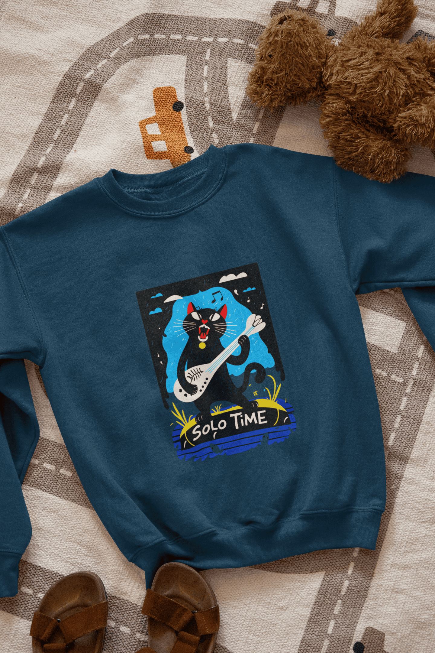 A navy blue sweatshirt with a design printed on the front lying down on the floor. The design contains a cat holding a fish bone like a guitar and appearing to play it aggressively. There's a text saying Solo Time at the bottom of the design.