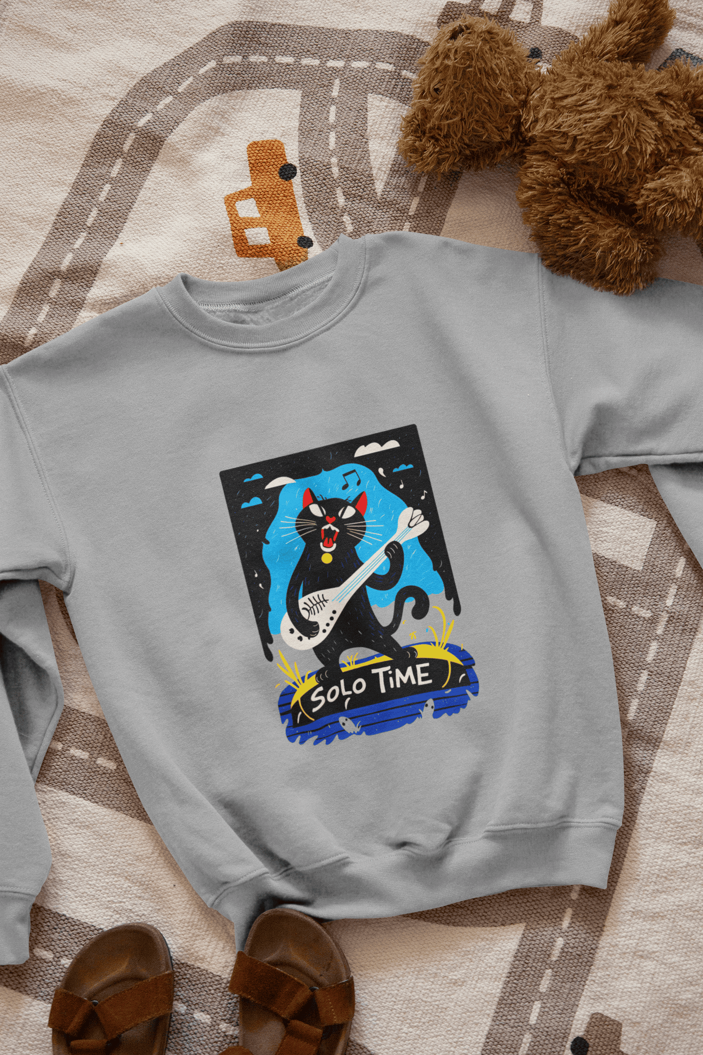 A grey sweatshirt with a design printed on the front lying down on the floor. The design contains a cat holding a fish bone like a guitar and appearing to play it aggressively. There's a text saying Solo Time at the bottom of the design.