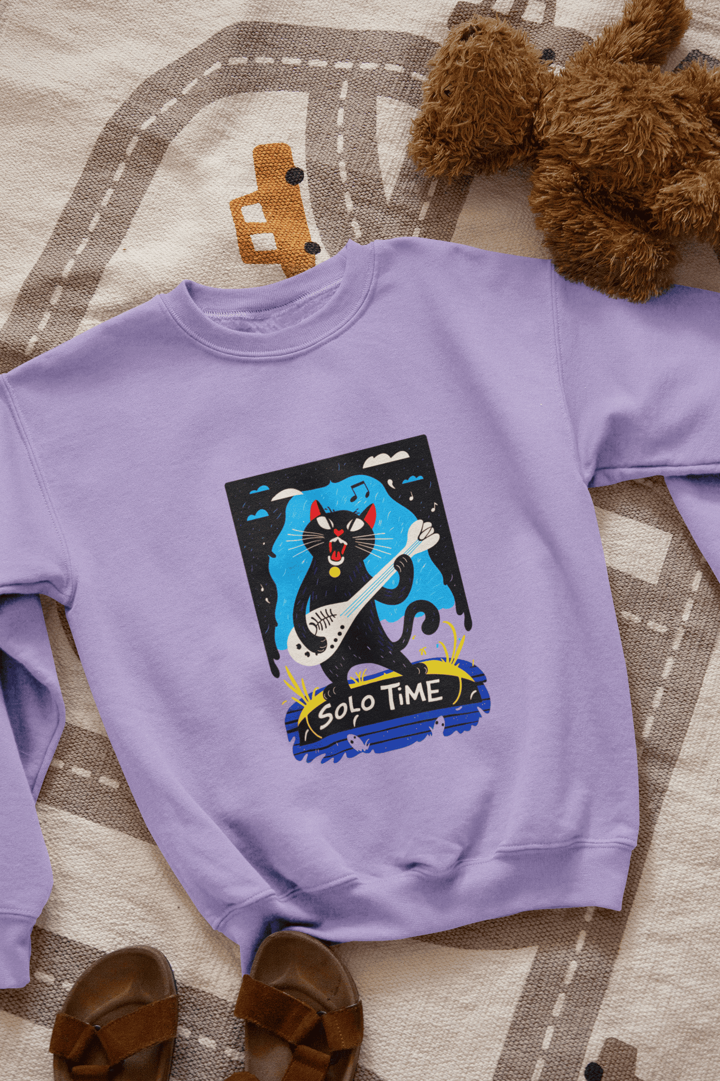 A lavender sweatshirt with a design printed on the front lying down on the floor. The design contains a cat holding a fish bone like a guitar and appearing to play it aggressively. There's a text saying Solo Time at the bottom of the design.