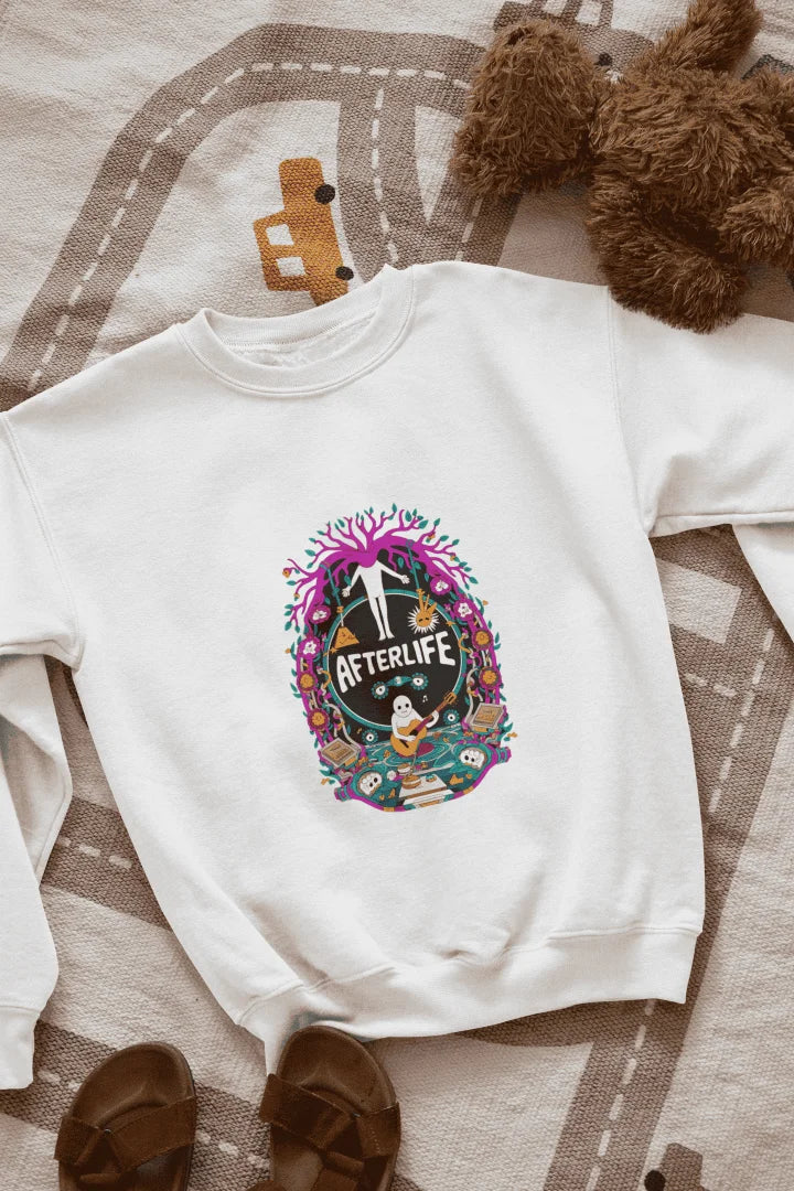 White sweatshirt featuring a colorful graphic design with a circular Afterlife text, showing a person with outstretched arms standing in front of a tree with pink and purple leaves. The design includes a light blue sky with white clouds, a person in a blue shirt smiling, and brown sandals and a teddy bear on a beige and white patterned blanket.