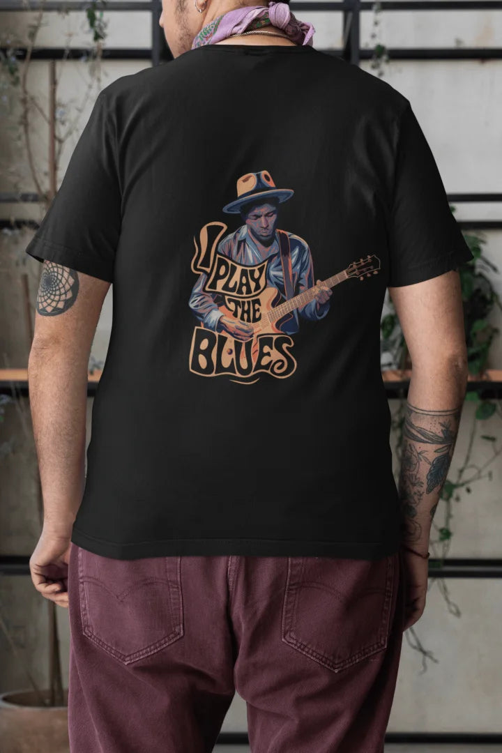 Man wearing a black back print oversized t-shirt with an illustration of a man playing a guitar and the words Play the Blues above him. The man is wearing a blue hat, standing in front of a brick wall with plants in the background.