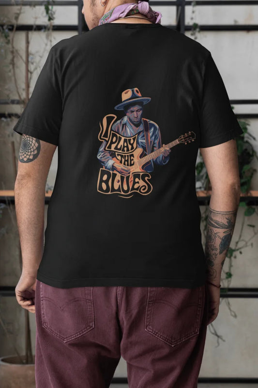 Man wearing a black back print oversized t-shirt with an illustration of a man playing a guitar and the words Play the Blues above him. The man is wearing a blue hat, standing in front of a brick wall with plants in the background.