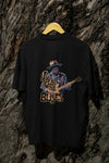 Black back print oversized t-shirt hanging on a wooden hanger against a dark background, featuring an illustration of a man in a blue shirt and cowboy hat playing an electric guitar with the words I Play the Blues in bold, cursive font above him. The design uses orange and yellow colors and has an 80s or 90s retro style.