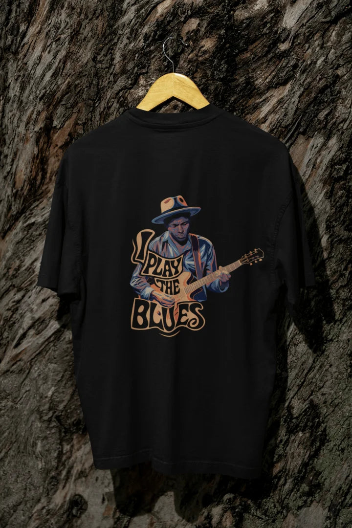 Black back print oversized t-shirt hanging on a wooden hanger against a dark background, featuring an illustration of a man in a blue shirt and cowboy hat playing an electric guitar with the words I Play the Blues in bold, cursive font above him. The design uses orange and yellow colors and has an 80s or 90s retro style.