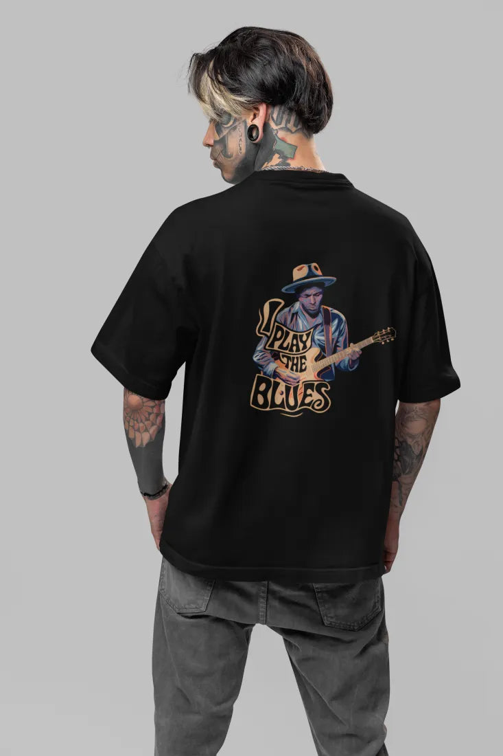 Man wearing a black back print oversized t-shirt with an illustration of a man playing a guitar and the words Play the Blues above him. The man is wearing a blue hat, standing in front of a brick wall with plants in the background.