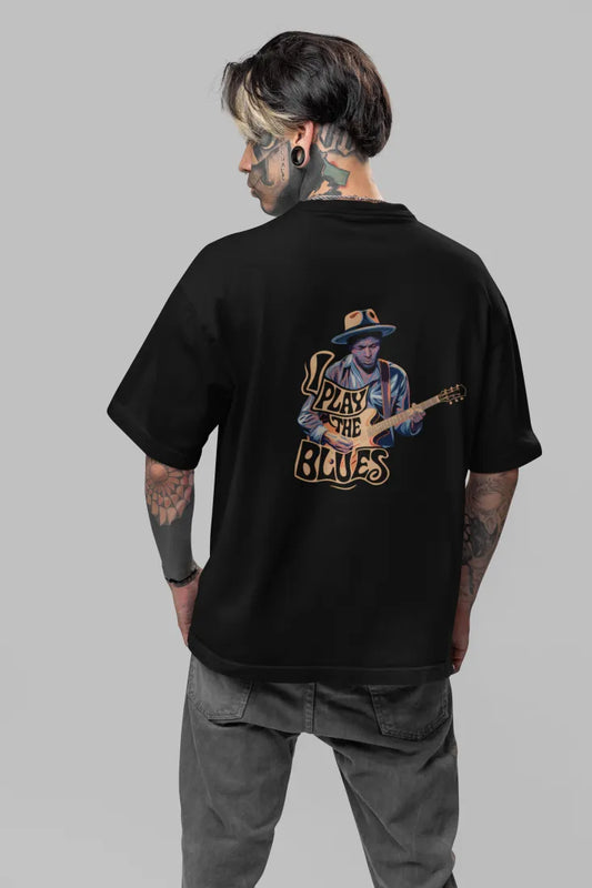 Man wearing a black back print oversized t-shirt with an illustration of a man playing a guitar and the words Play the Blues above him. The man is wearing a blue hat, standing in front of a brick wall with plants in the background.