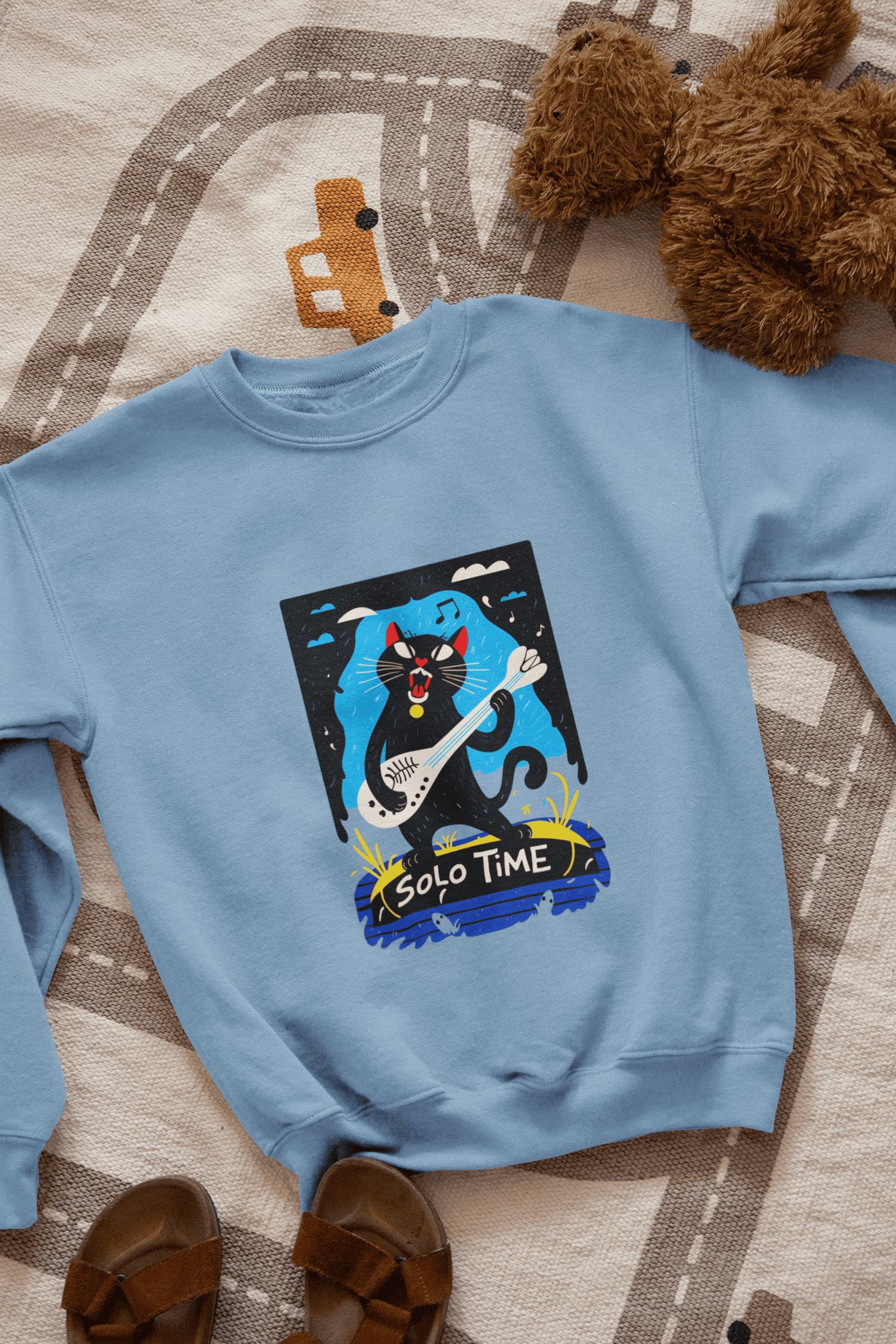 A baby blue sweatshirt with a design printed on the front lying down on the floor. The design contains a cat holding a fish bone like a guitar and appearing to play it aggressively. There's a text saying Solo Time at the bottom of the design.