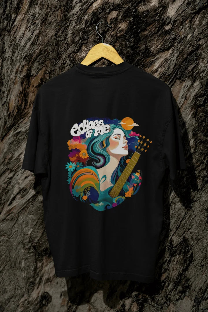 A black oversized T-shirt with a vibrant back graphic featuring a woman with teal-and-purple hair playing a golden guitar. Surrounded by colorful flowers, swirling patterns, and a crescent moon, the text “Echoes of Me” is integrated. The design has a 1970s-inspired psychedelic style, evoking a dreamy, musical vibe.