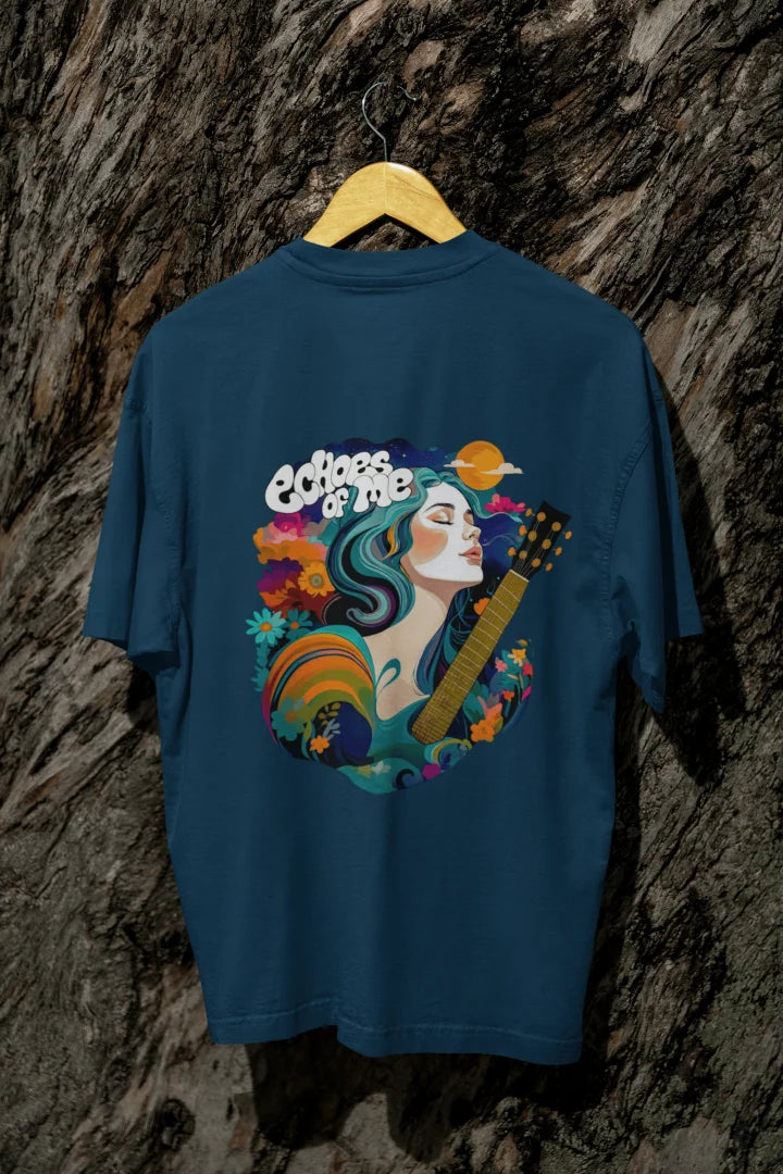 A navy blue T-shirt with a vibrant back graphic featuring a woman with teal-and-purple hair playing a golden guitar. Surrounded by colorful flowers, swirling patterns, and a crescent moon, the text “Echoes of Me” is integrated. The design has a 1970s-inspired psychedelic style, evoking a dreamy, musical vibe.