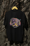 A black back printed oversized t-shirt with a vibrant psychedelic graphic of a man playing an electric guitar, surrounded by wavy pastel shapes. Bold text below reads “F**K THE SYSTEM.”