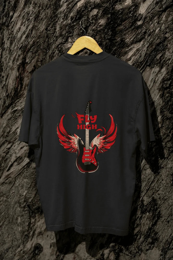 A black back printed oversized t-shirt with a graphic of an electric guitar with red, outstretched wings. The words “FLY HIGH” appear above in bold font. The shirt hangs on a wooden hanger against dark-grey tree bark.