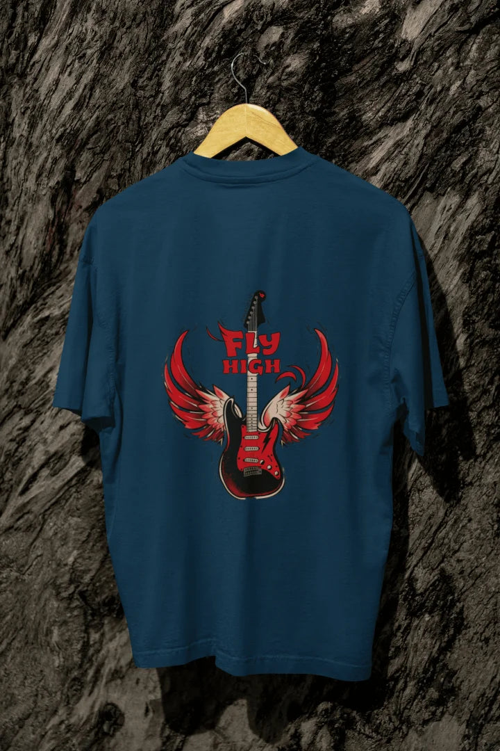 A navy blue back printed oversized t-shirt with a graphic of an electric guitar with red, outstretched wings. The words “FLY HIGH” appear above in bold font. The shirt hangs on a wooden hanger against dark-grey tree bark.