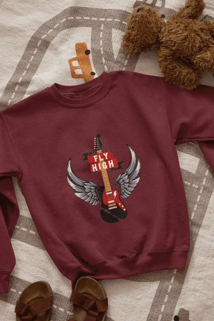 A maroon sweatshirt with a graphic design of a black electric guitar with gray and white angel wings, and a red “FLY HIGH” banner. The sweatshirt is laid flat on a light beige road-patterned rug, with brown leather sandals at the bottom left. Soft lighting highlights the graphic, creating a relaxed, casual atmosphere.