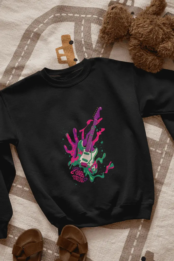 A black sweatshirt kept on the floor containing a psychedelic design pattern. The design contains of guitar made up of paint splashes, and a hand at the back, trying to grab it There is a text saying let me grab that neck.