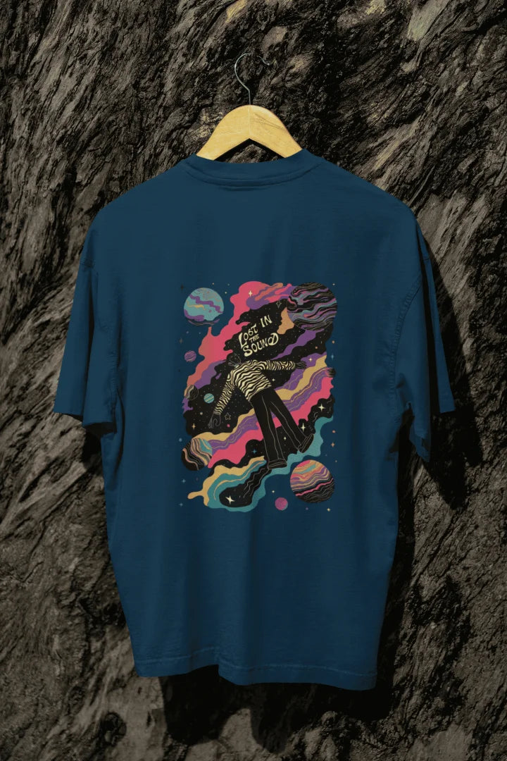 A navy blue oversized T-shirt hanging against a tree trunk. The T-shirt contains a back print of a man floating in space. There’s a text saying Lost in the Sound