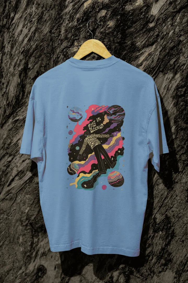 A baby blue oversized T-shirt hanging against a tree trunk. The T-shirt contains a back print of a man floating in space. There’s a text saying Lost in the Sound