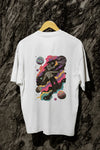 A white oversized T-shirt hanging against a tree trunk. The T-shirt contains a back print of a man floating in space. There’s a text saying Lost in the Sound