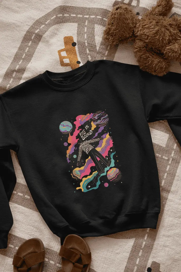 A printed black sweatshirt lying on the floor. The design of the sweatshirt contains of a man floating in space. The design is a colourful design with tiny planets and stars. There’s a text saying Lost in the sound.