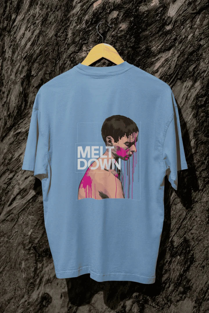 A baby blue oversized T-shirt hanging against a tree, containing the design of a man in grief. The man is covered in pink paint splashes with paint dripping down from his face. There’s a text saying Meltdown on the design. 