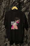 A black oversized T-shirt hanging against a tree, containing the design of a man in grief. The man is covered in pink paint splashes with paint dripping down from his face. There’s a text saying Meltdown on the design. 