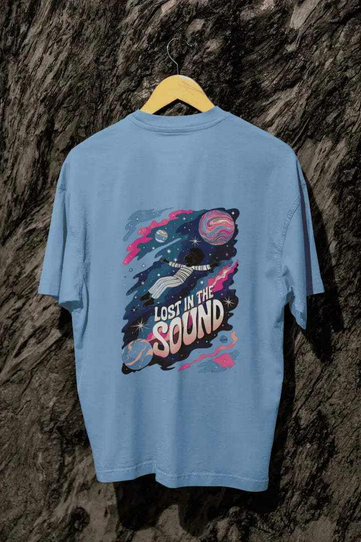 A baby blue oversized t-shirt with a back print hanging on a tree trunk. The print design contains a kid floating in space with planets and starts surrounding him. There is a text saying Lost In The Sound on the design.