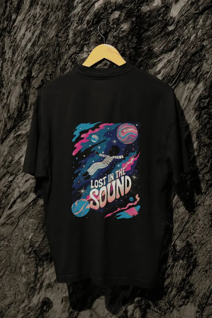 A black oversized t-shirt with a back print hanging on a tree trunk. The print design contains a kid floating in space with planets and starts surrounding him. There is a text saying Lost In The Sound on the design.
