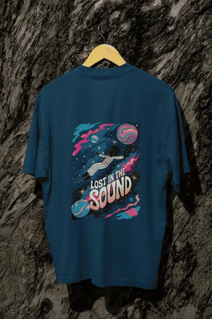 A navy blue oversized t-shirt with a back print hanging on a tree trunk. The print design contains a kid floating in space with planets and starts surrounding him. There is a text saying Lost In The Sound on the design.