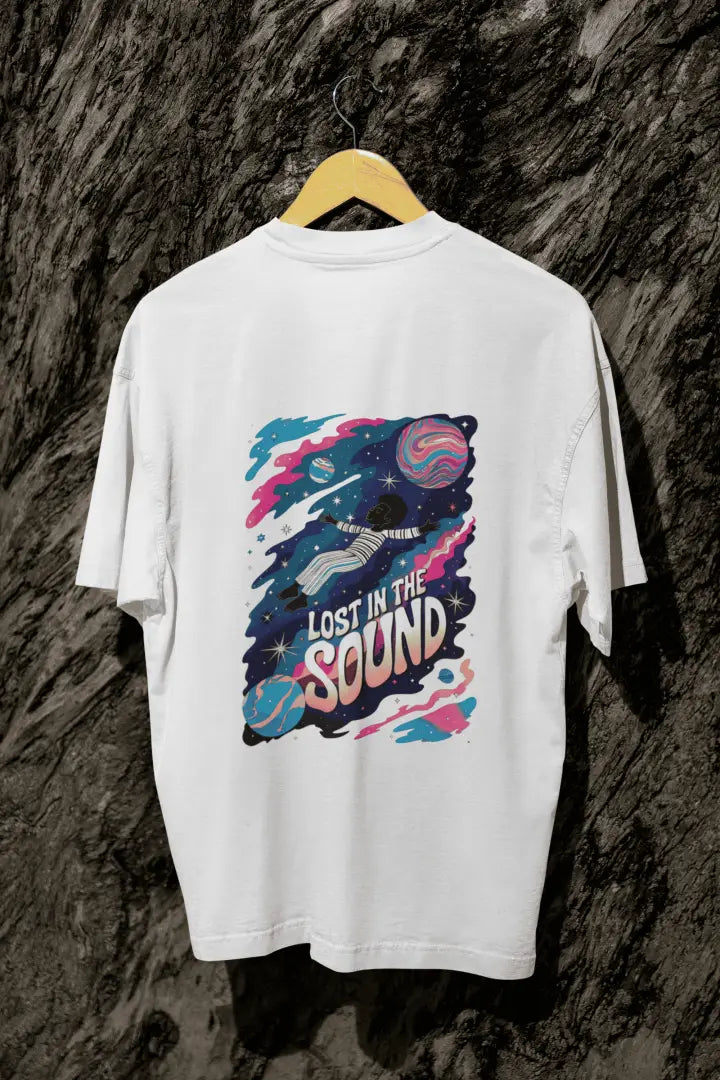 A white oversized t-shirt with a back print hanging on a tree trunk. The print design contains a kid floating in space with planets and starts surrounding him. There is a text saying Lost In The Sound on the design.