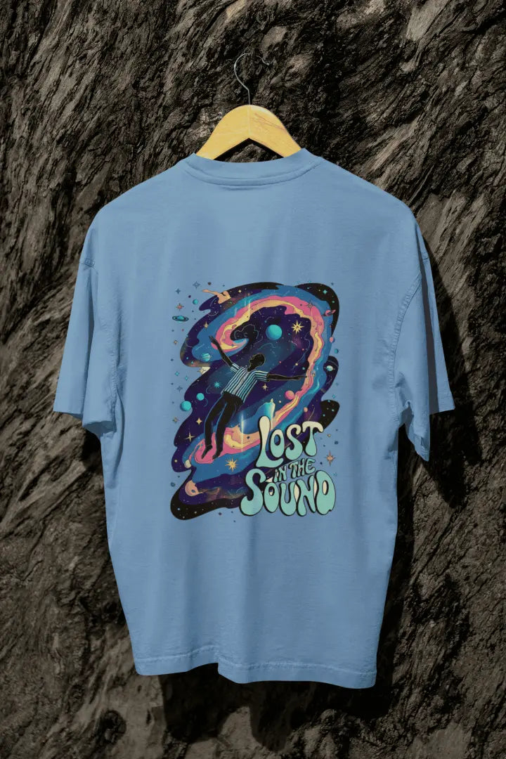 A baby blue back printed oversized t-shirt with a back print hanging on a tree trunk. The back print contains a man floating in space with planets and stars around him. There's a text saying Lost in The Sound on the bottom right of the design.