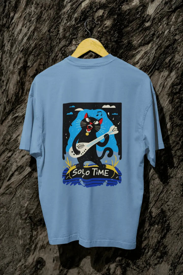 A baby blue back printed oversized t-shirt with a back print hanging from a tree trunk. The design contains a cat holding a fish bone like a guitar and appears to play it aggressively. There's a text saying Solo Time at the bottom of the design.