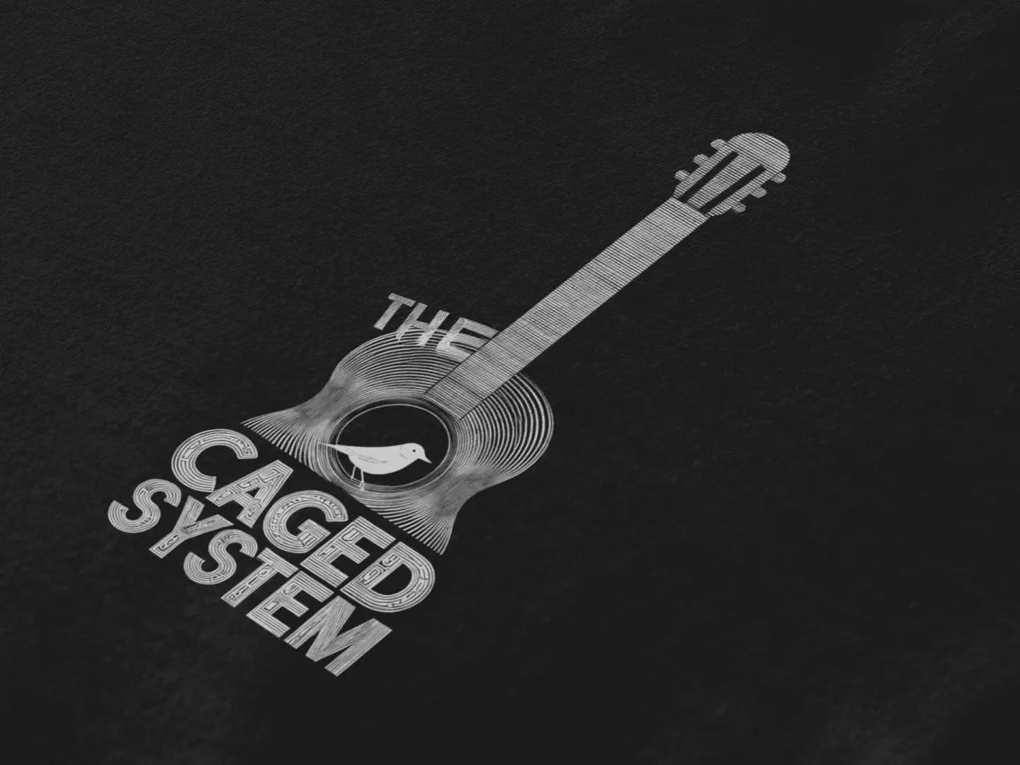 Close up of a t-shirt with a minimal design printed on the front. The design consists of an acoustic guitar with a bird sitting inside it. There's a text saying The Caged System at the bottom of the design.